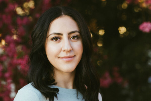 A woman with dark hair and a grey dress looks in to the camera with a smile